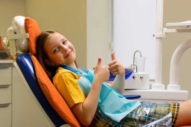 A cheerful schoolgirl gives a thumbs-up while sitting in a dental chair after a routine dentist appointment